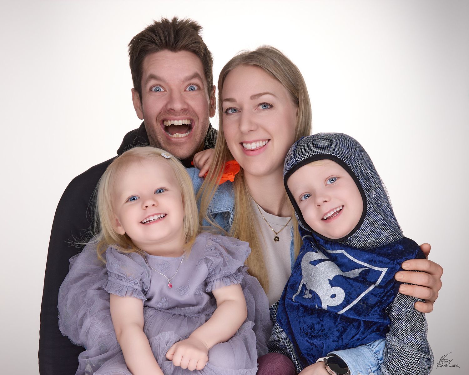 Smiling family portrait of a playful man, a blonde woman, a toddler girl in a gray dress and a young boy in a hooded blue top.