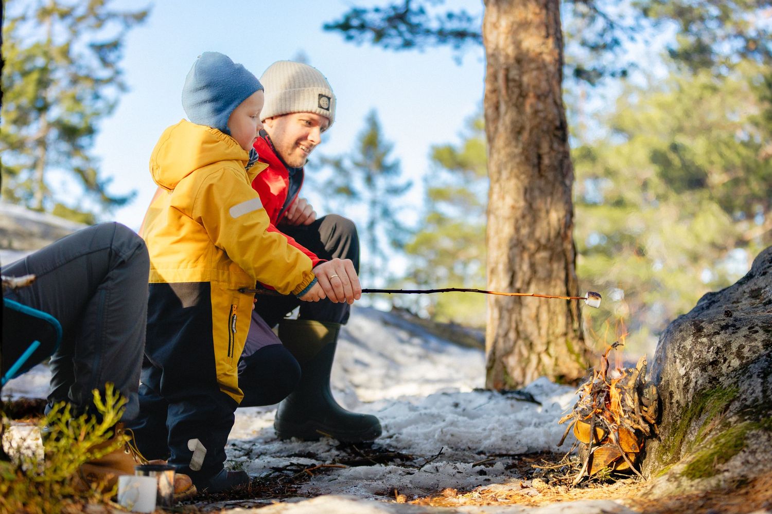 Two people in warm winter jackets and beanies crouch beside a snowy tree in a pine forest, roasting food over a small campfire.