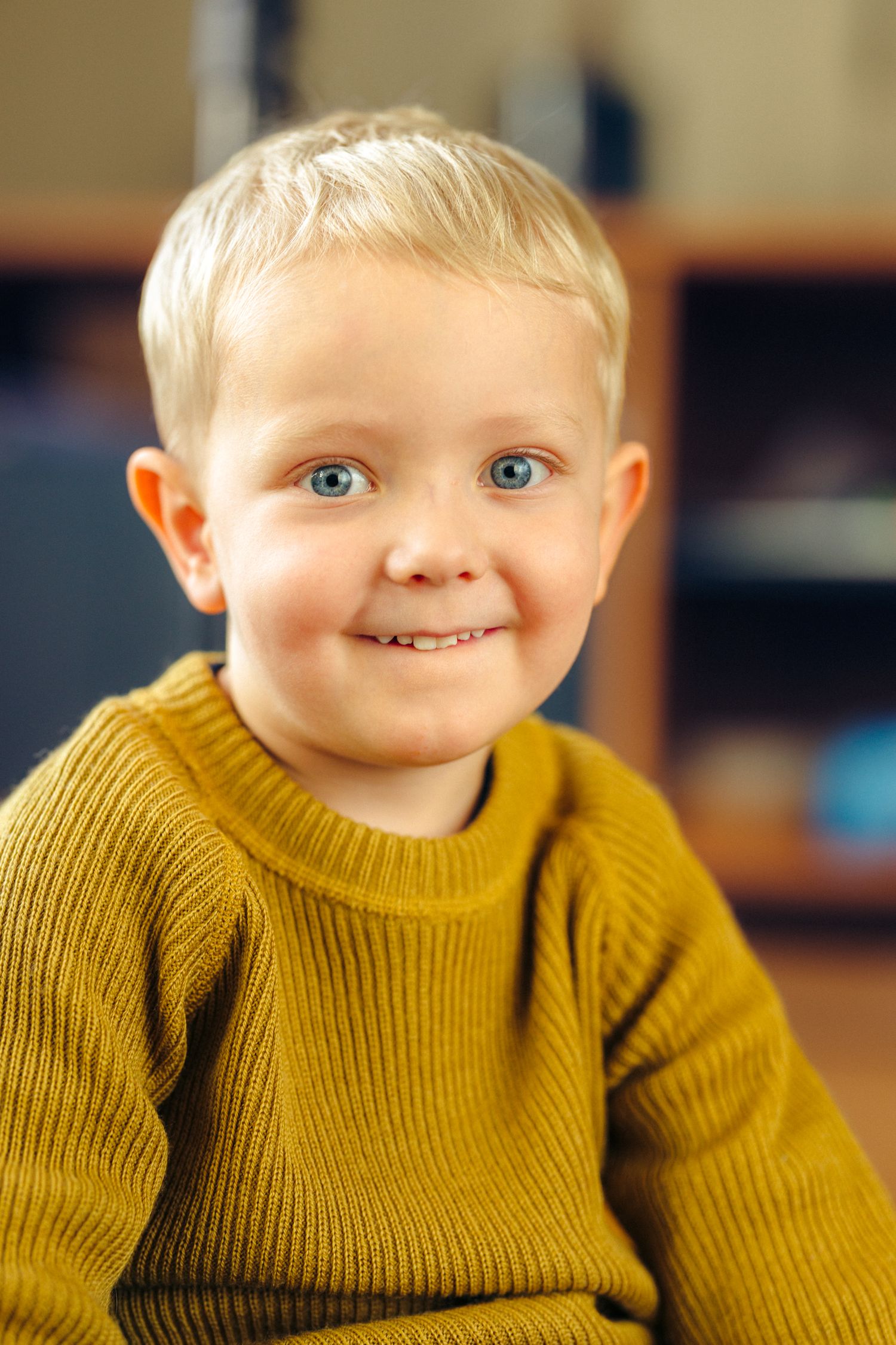 Smiling toddler with short blond hair and blue eyes wearing a mustard yellow knit sweater, seated indoors in a softly blurred room.