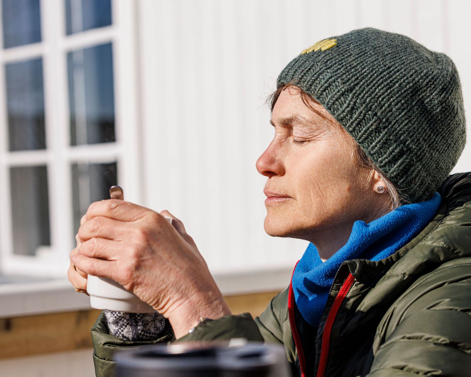 Person wearing a green winter coat and beanie hat takes a sip from a steaming beverage outdoors.