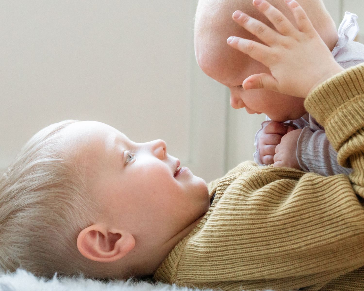 A young child in a mustard knit sweater lies on a soft rug and gently holds a newborn baby up in the air.