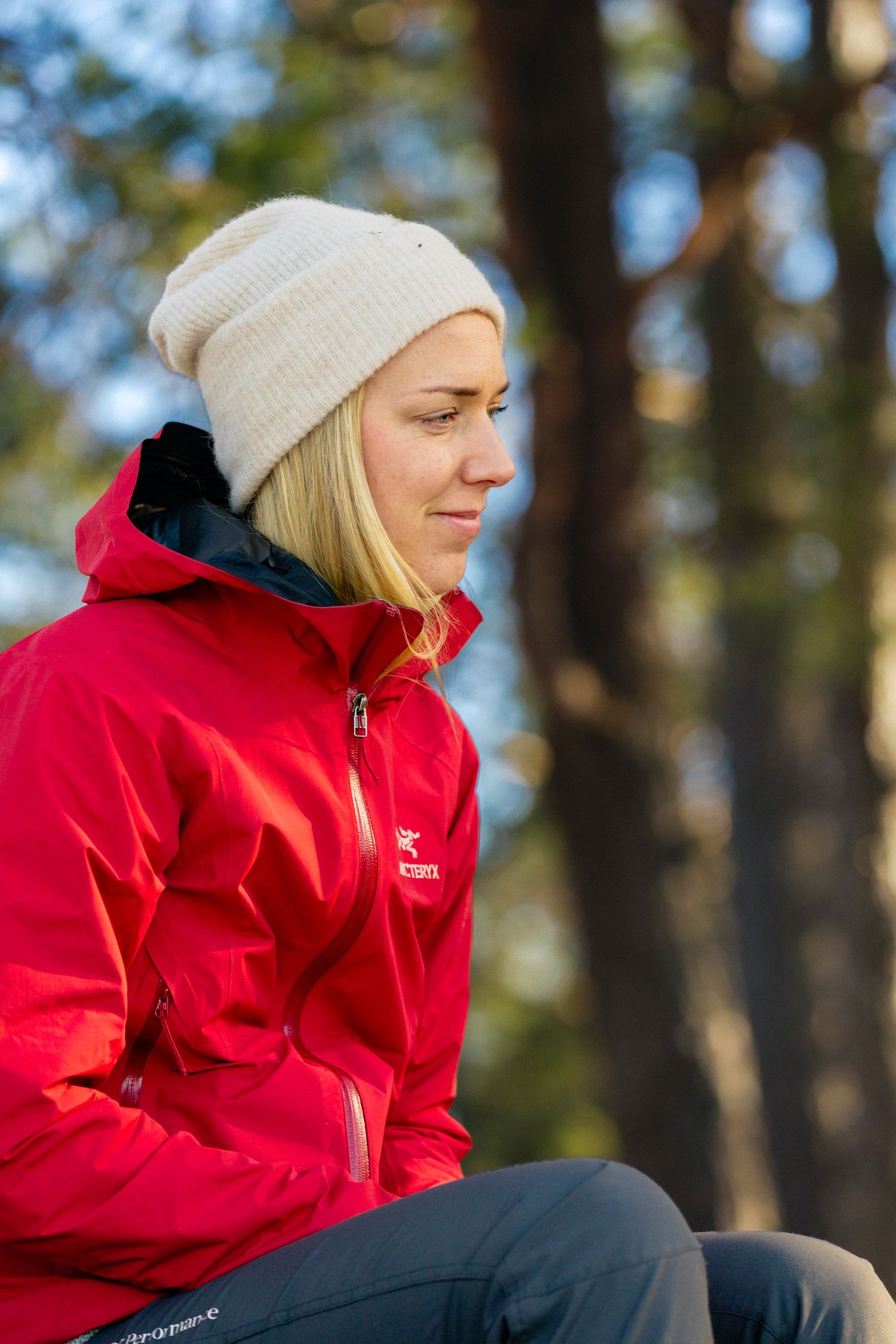 Woman in a red hooded jacket and cream knit beanie sits outdoors in profile, surrounded by blurred forest trees in soft sunlight.