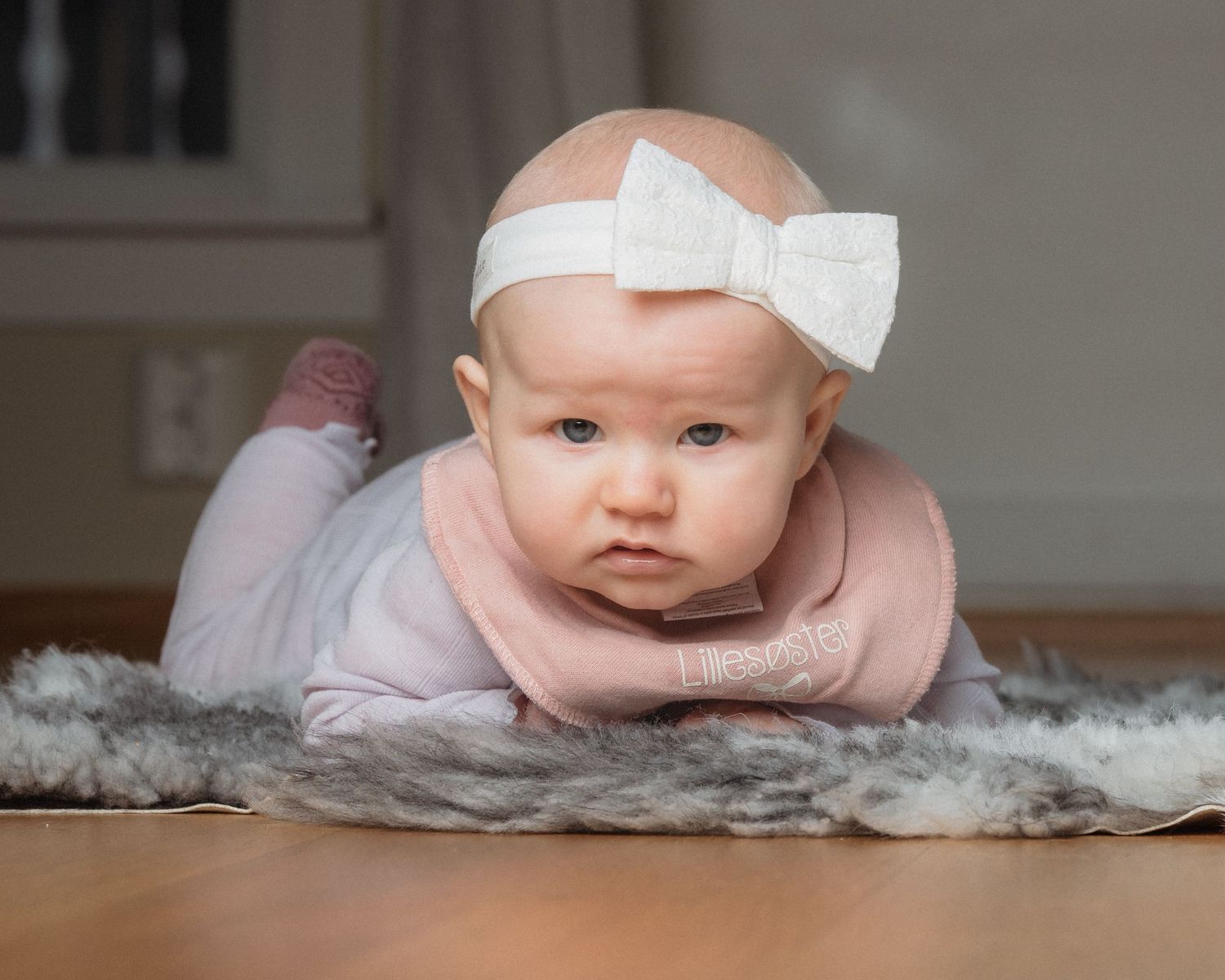 Baby in a white bow headband and pink bib lies on a gray faux fur rug on a wooden floor, looking at the camera.
