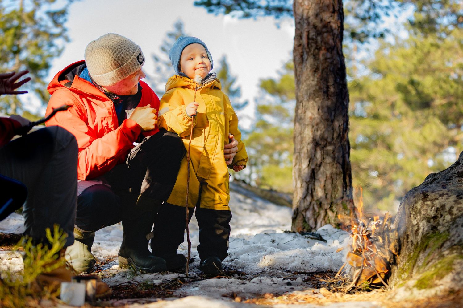 Adult in an orange jacket and a child in a yellow coat and blue hat sit by a small campfire in a sunny, snowy pine forest.