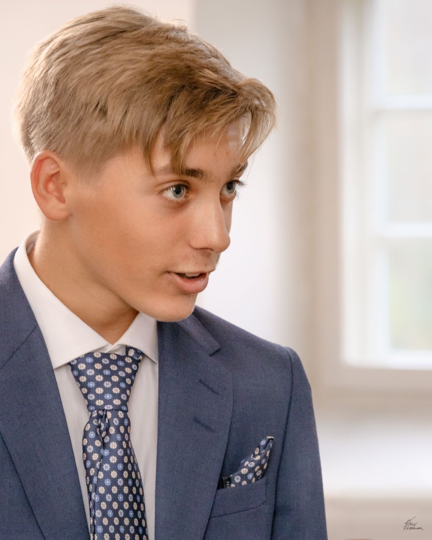 Portrait of young person in grey suit and patterned tie looking to the side in bright interior.