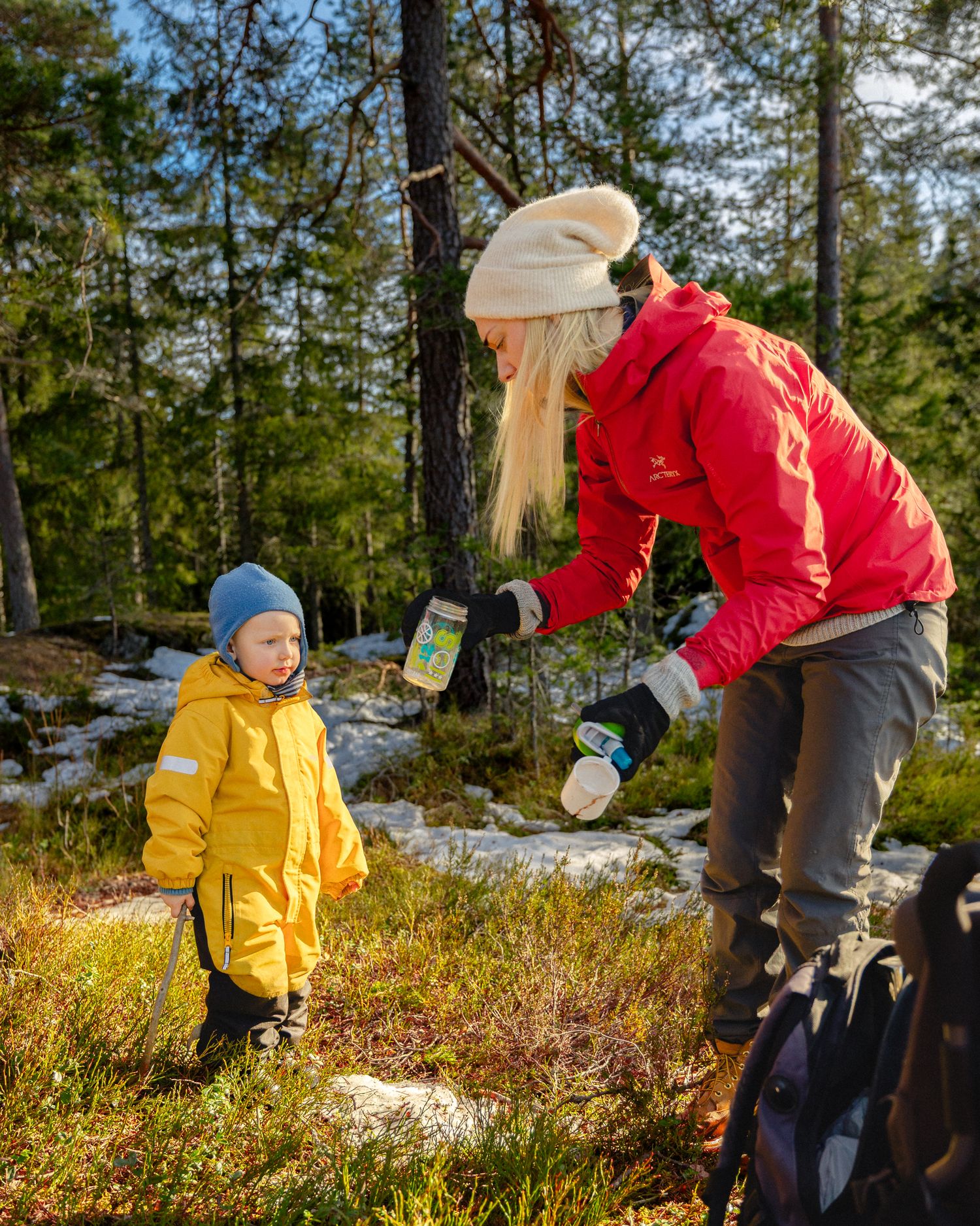 Woman in a red jacket and white beanie hands a small bottle to a toddler in a yellow rain suit and blue hat in a sunny autumn forest.