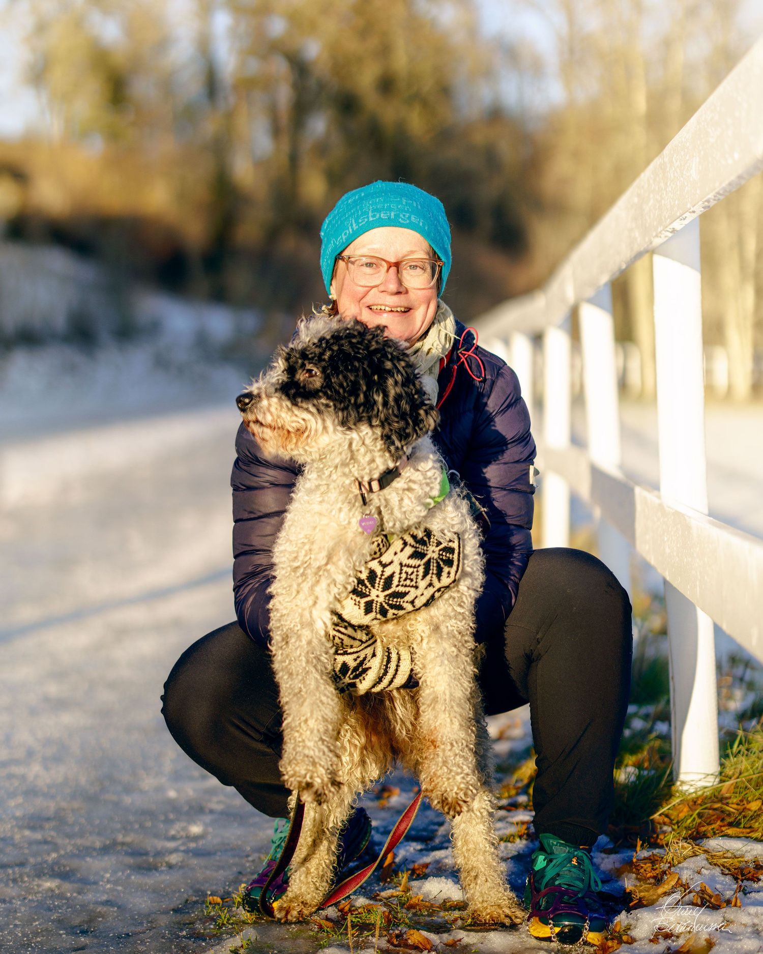 A person sits on a snowy path with a fluffy white dog on a leash during a winter day.