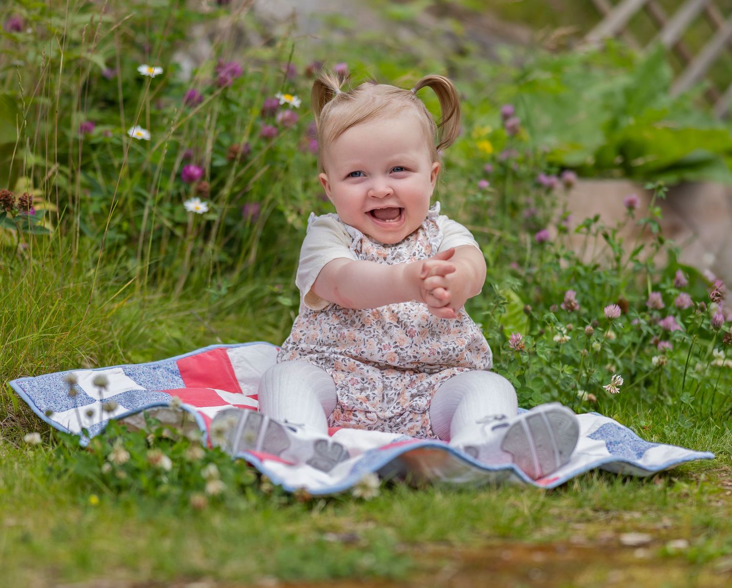 Baby sitting in grass with flowers and blankets during outdoor photo session.