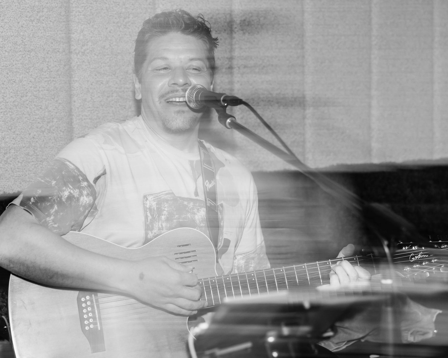 A musician smiling while performing with an acoustic guitar in a black and white photograph.