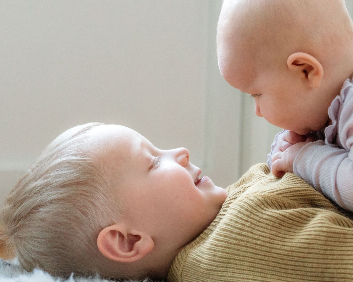 Two babies face each other up close, one lying on its back smiling at the other, who leans in curiously over a cozy knit sweater.