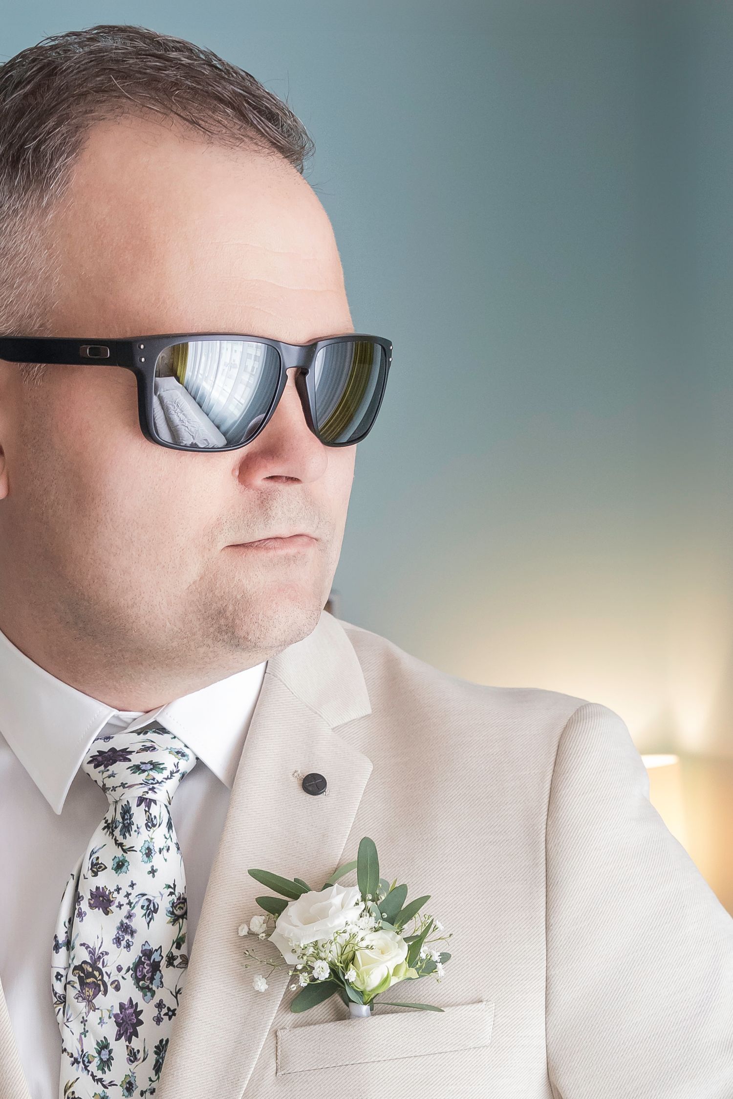 Close-up profile view of someone wearing a white suit jacket, floral tie, and sunglasses against a soft blurred background.
