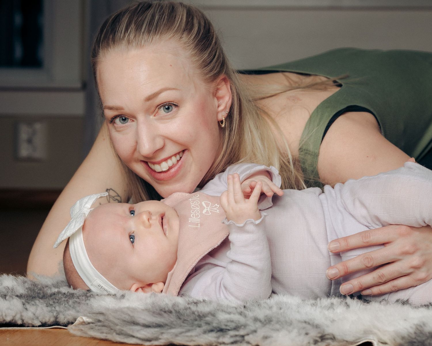 Smiling blonde mother lies on a soft fur rug beside her baby in a pink outfit and white headband, who gazes sideways at the camera.