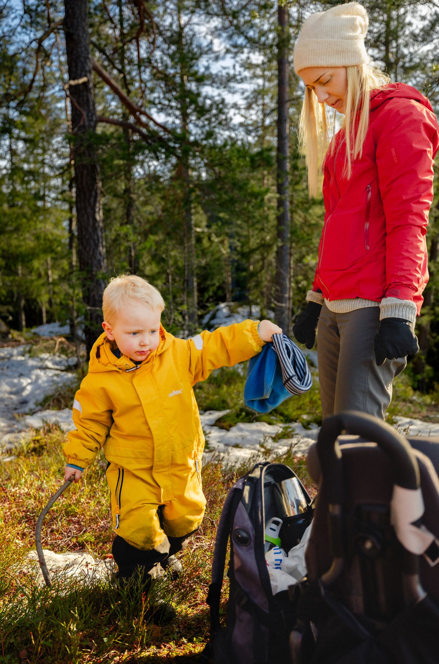 Toddler in a yellow rain suit holds a blue cloth beside a woman in a red jacket in a sunny forest with a backpack and carrier nearby.