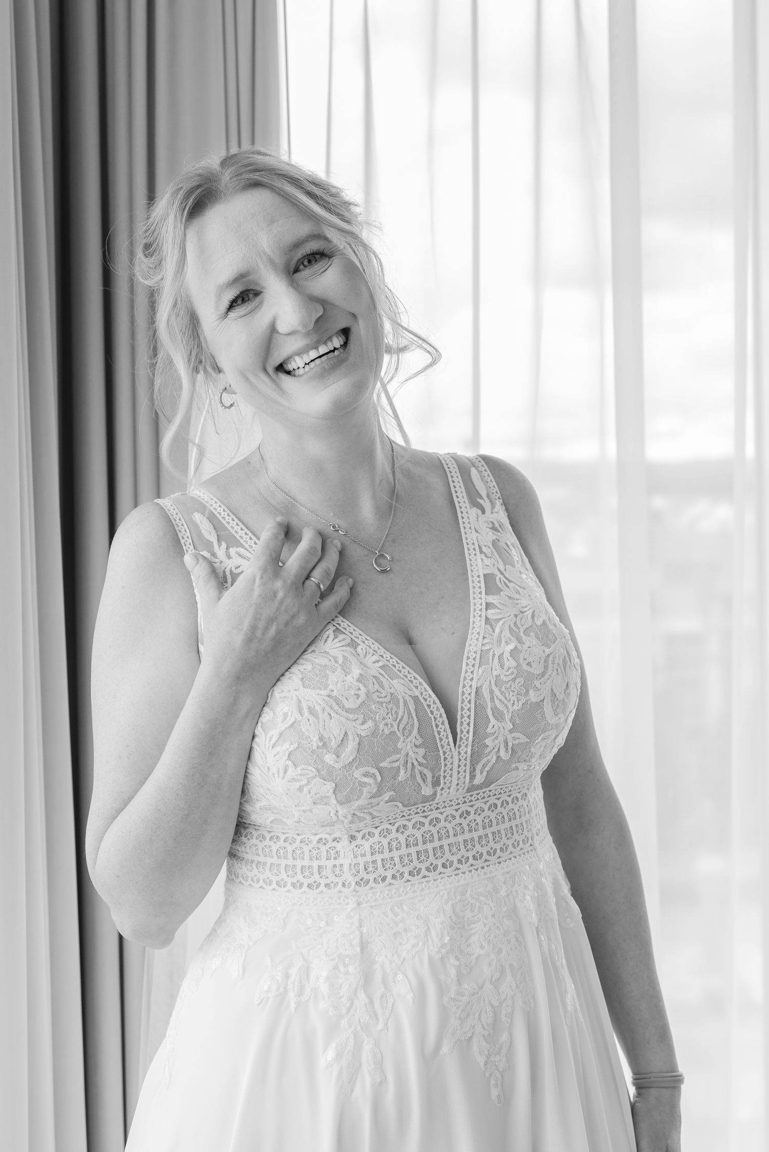 Joyful bride wearing a white lace wedding dress poses by window with natural light.