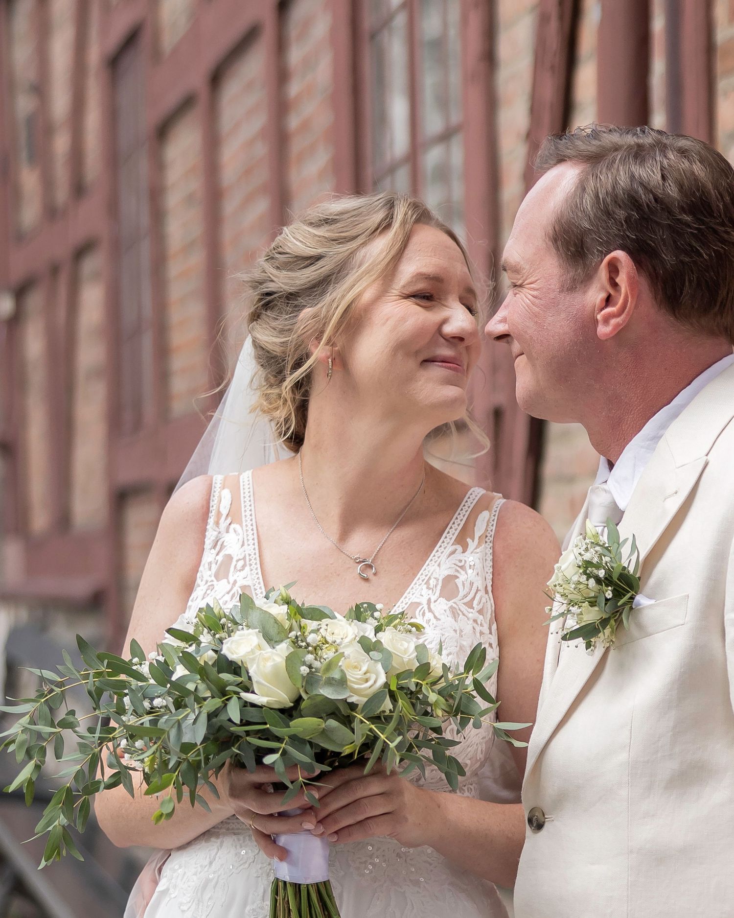 Romantic wedding couple sharing intimate moments against brick wall with white floral bouquet.