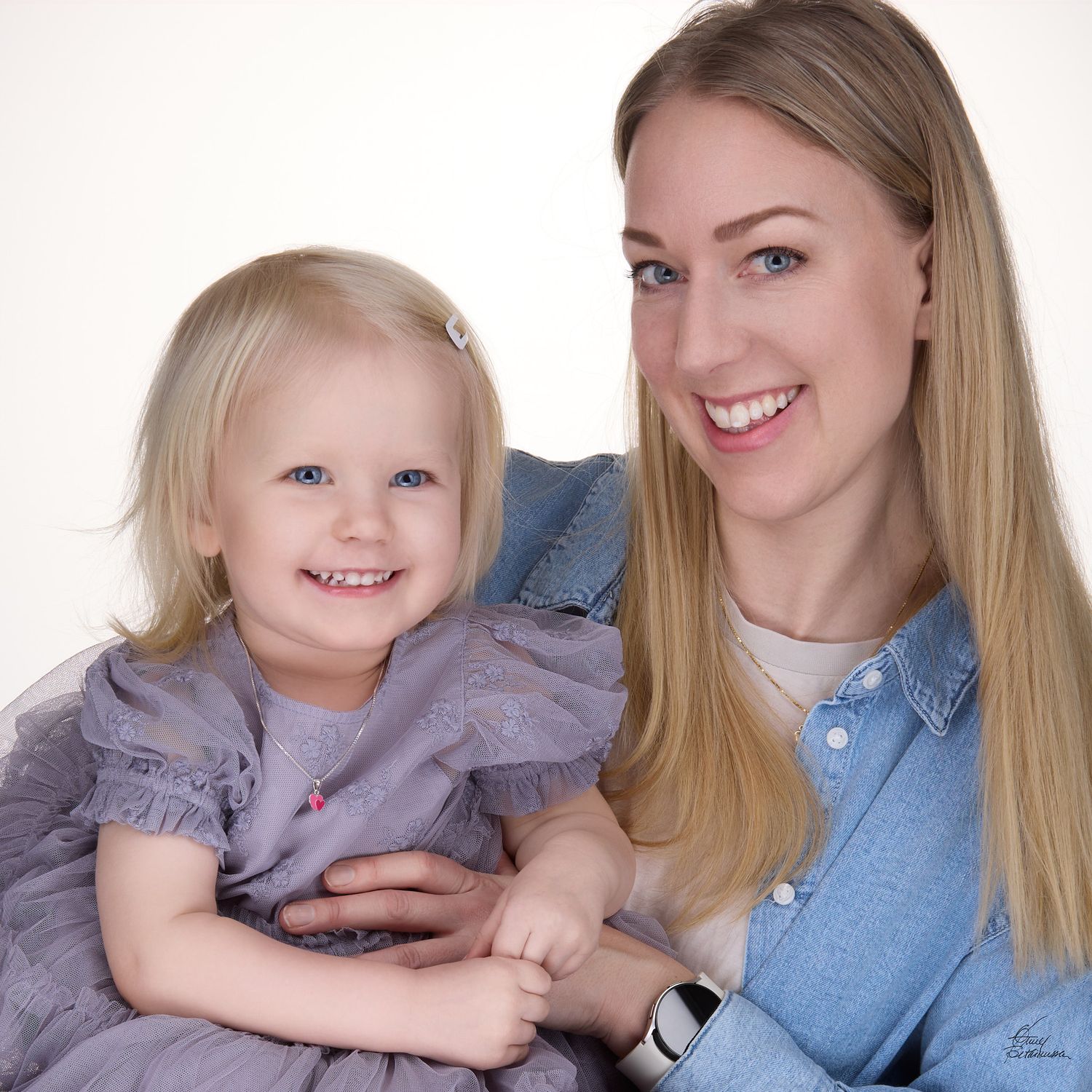 Smiling blonde woman in a denim shirt holding a laughing toddler girl in a gray ruffled dress in a bright studio portrait.