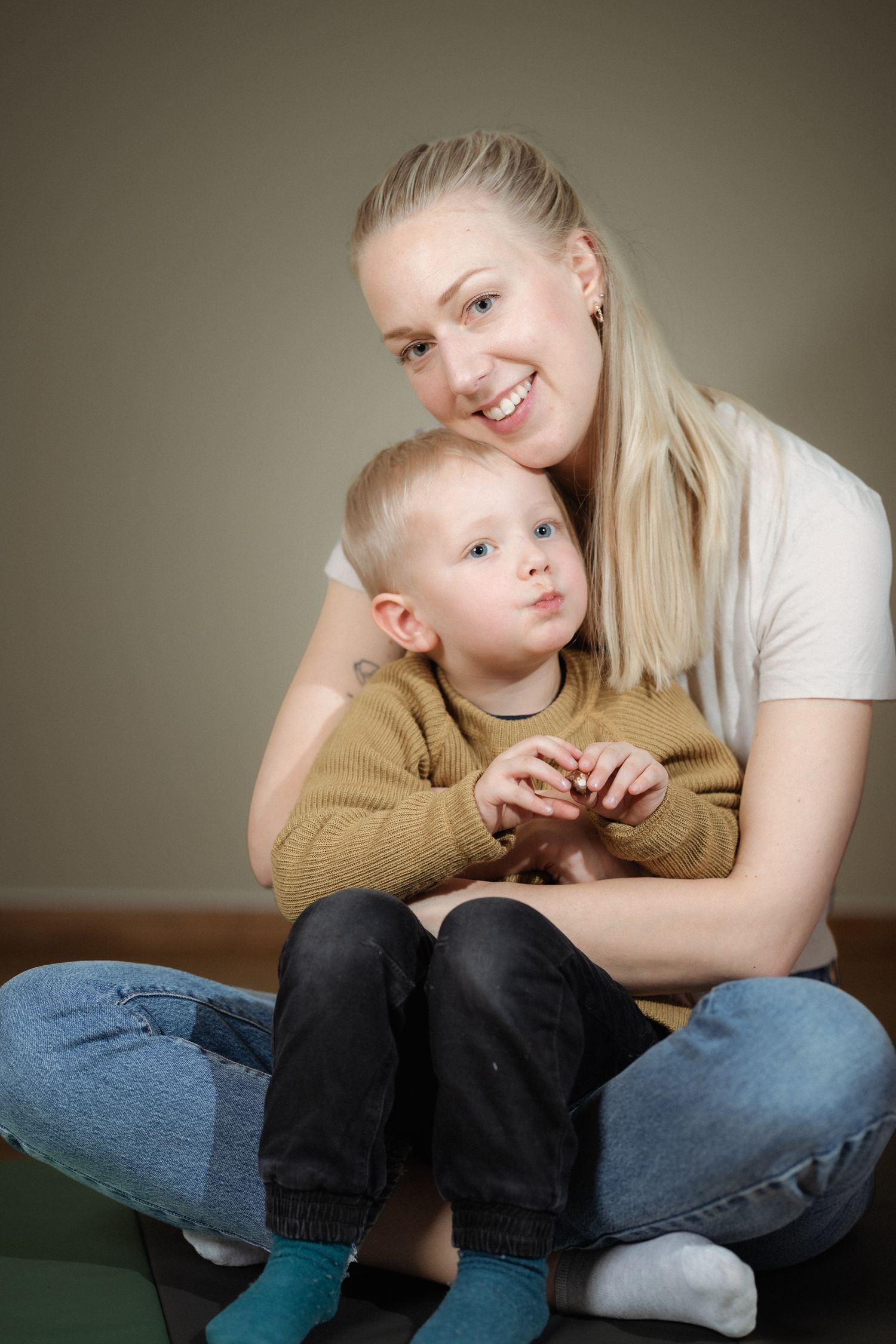 Smiling blonde woman in a white t-shirt and jeans sitting cross-legged and hugging a toddler in a mustard sweater and teal socks.