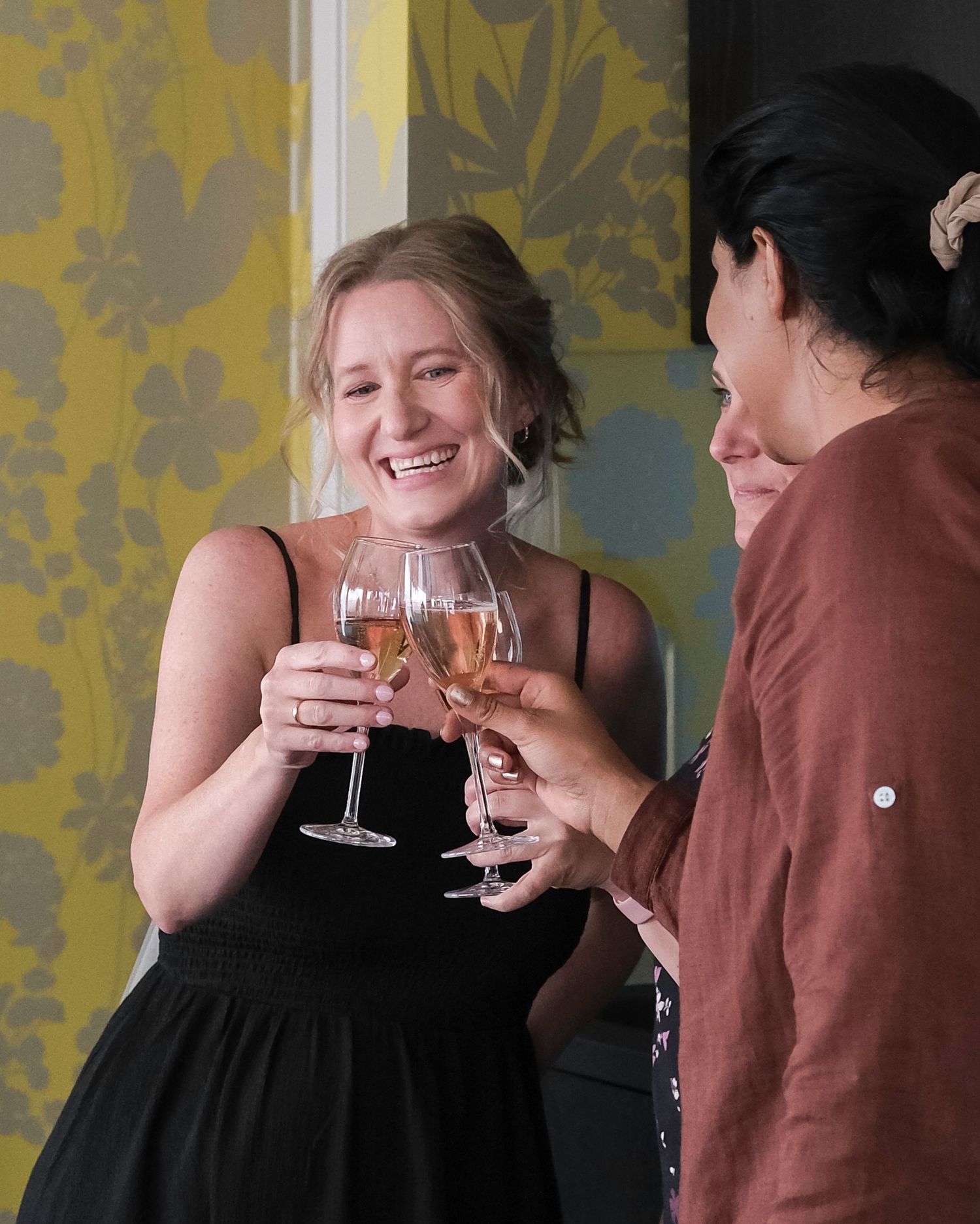 Two friends sharing laughter and toasting with wine glasses against a yellow floral wallpaper background.