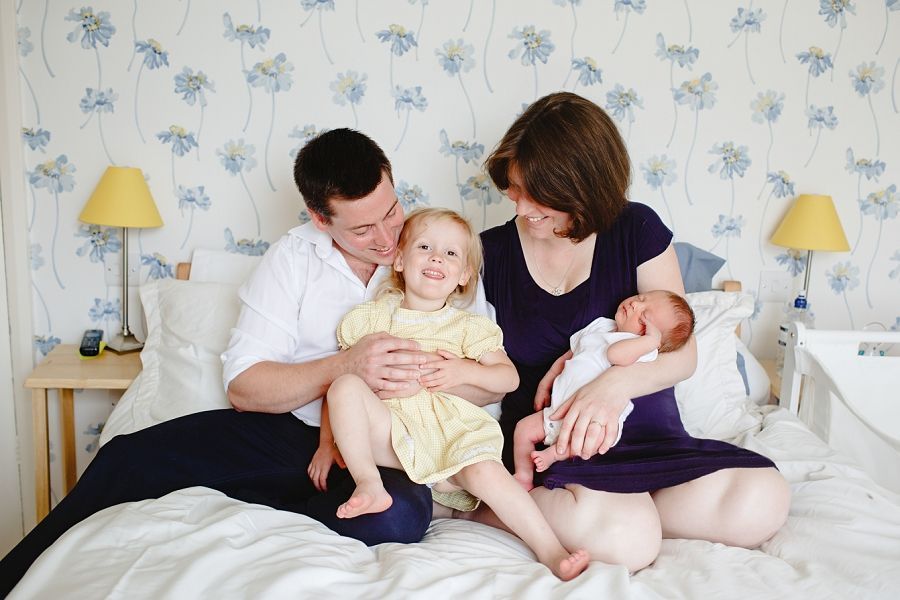 Family cuddles together on white bedding with blue floral wallpaper in background.