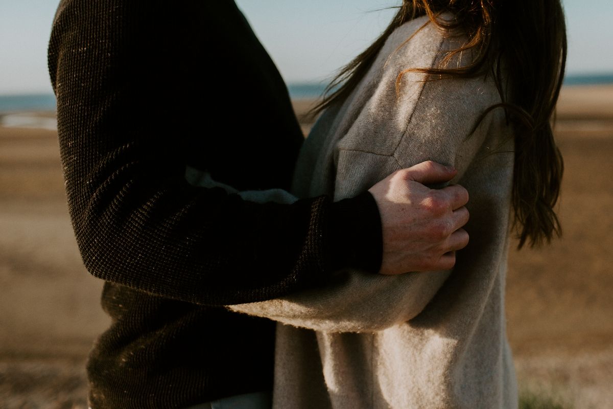 Couple embracing tenderly on a beach, woman in cream shawl held from behind by partner in dark sweater.