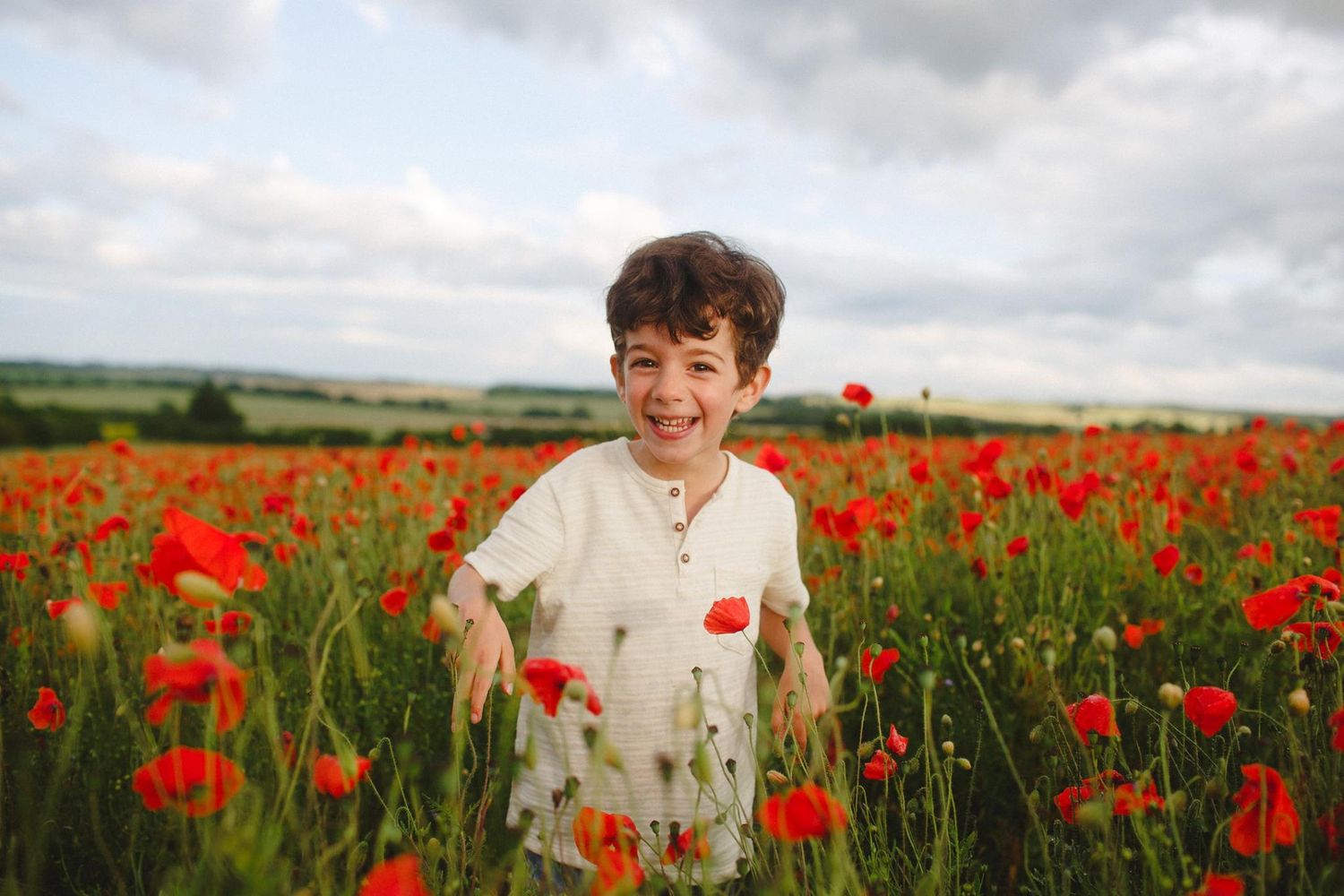 Child standing in a vibrant red poppy field under cloudy skies.