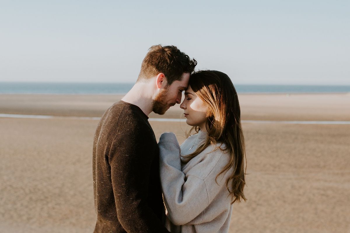 A romantic couple sharing an intimate moment on a sandy beach, touching foreheads tenderly with the ocean in the background.