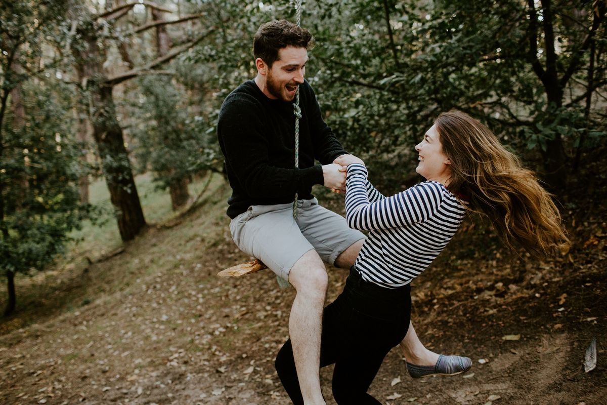 A joyful couple laughs and plays on a forest trail, with the man lifting the woman in a striped shirt.