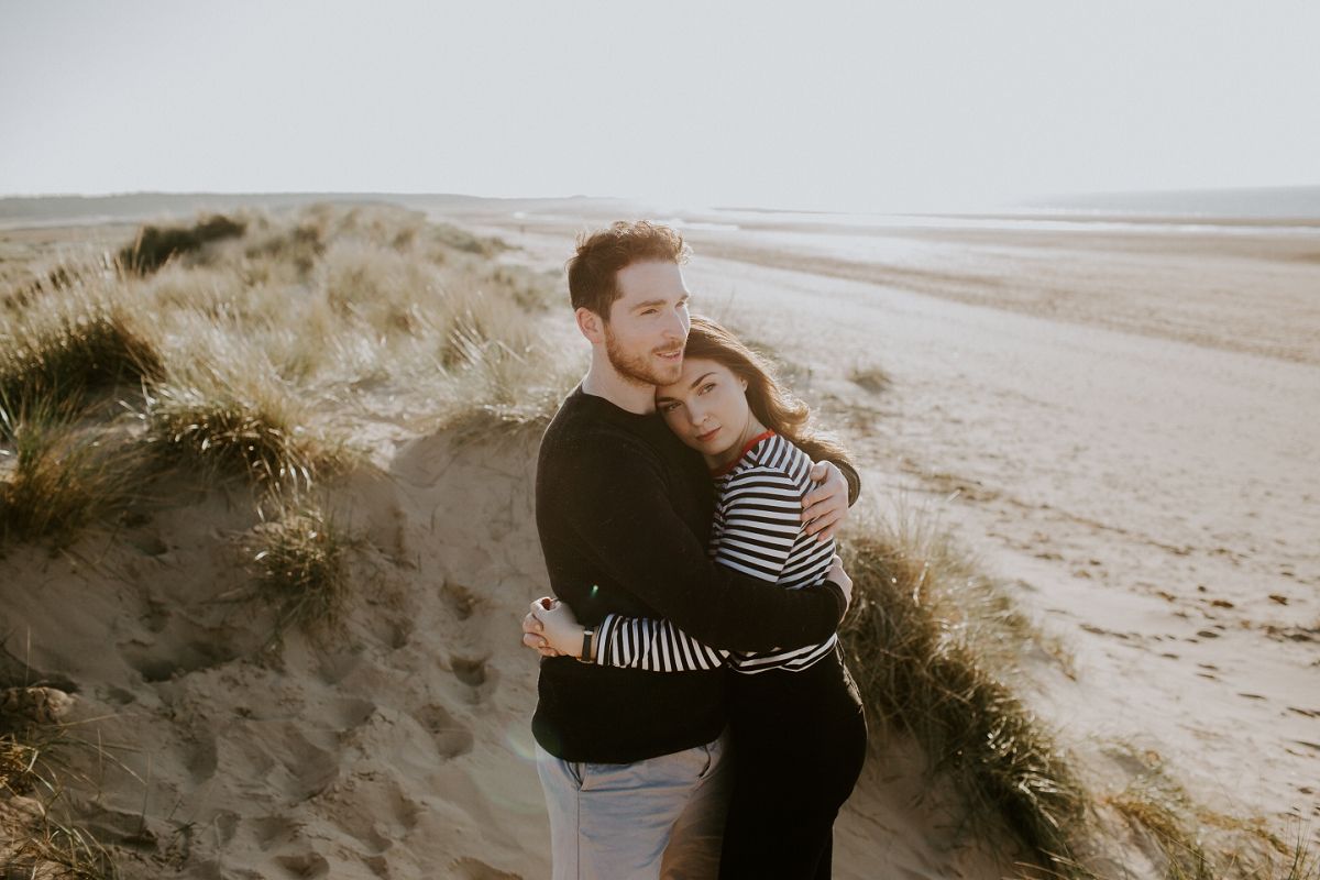 Couple embracing on sandy beach dunes, woman in striped top leaning on man in black sweater, serene coastal backdrop.
