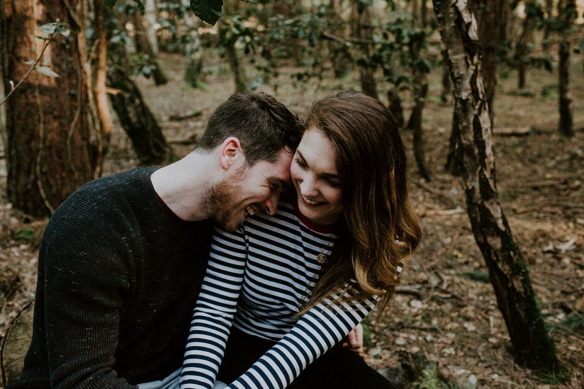 A couple sharing a tender moment together in a woodland setting, smiling and embracing among trees.