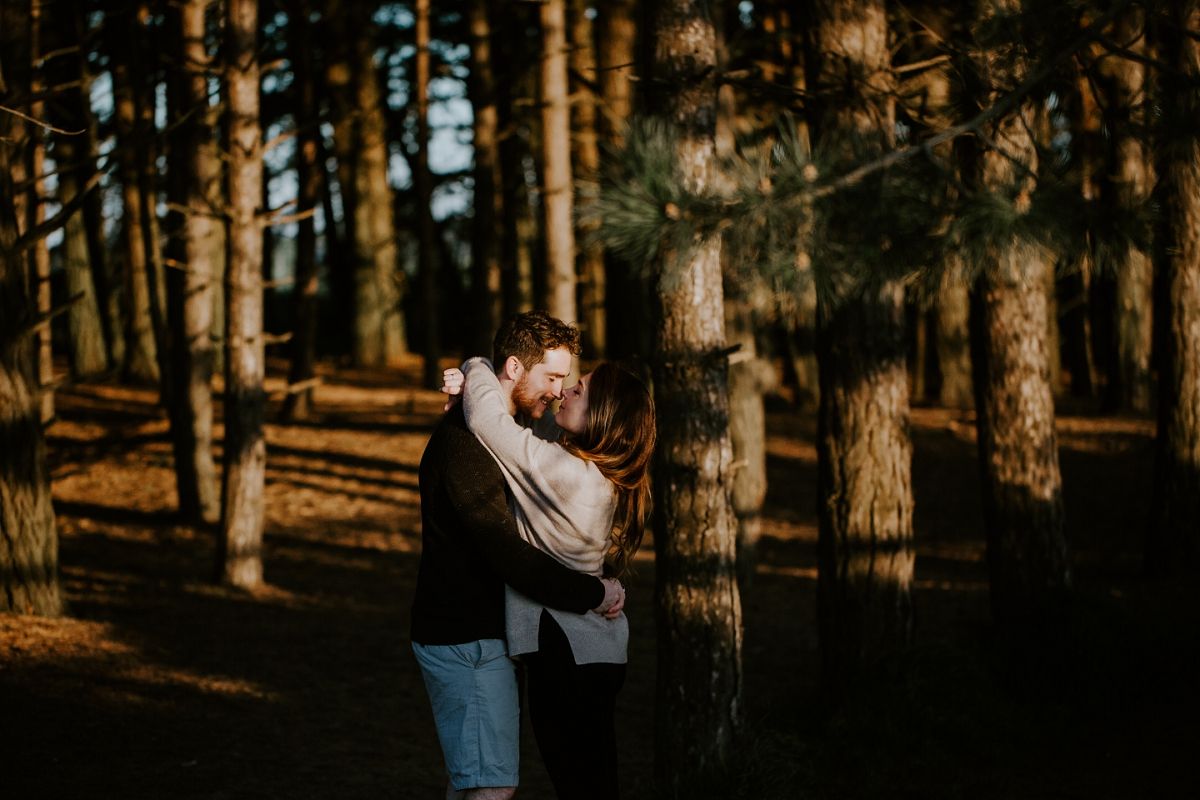 Couple embracing romantically among tall pine trees in golden hour sunlight during an outdoor engagement photo session.