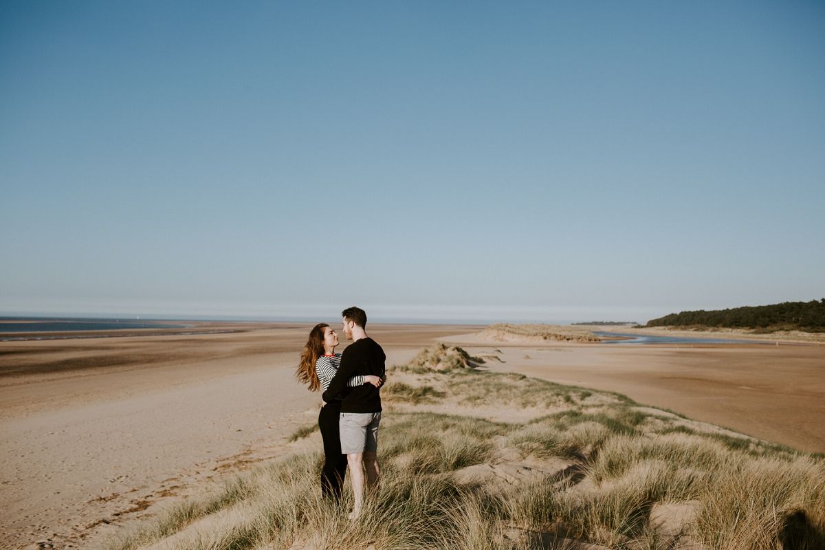 Couple embracing and kissing on sandy beach dunes with vast shoreline and clear blue sky backdrop.