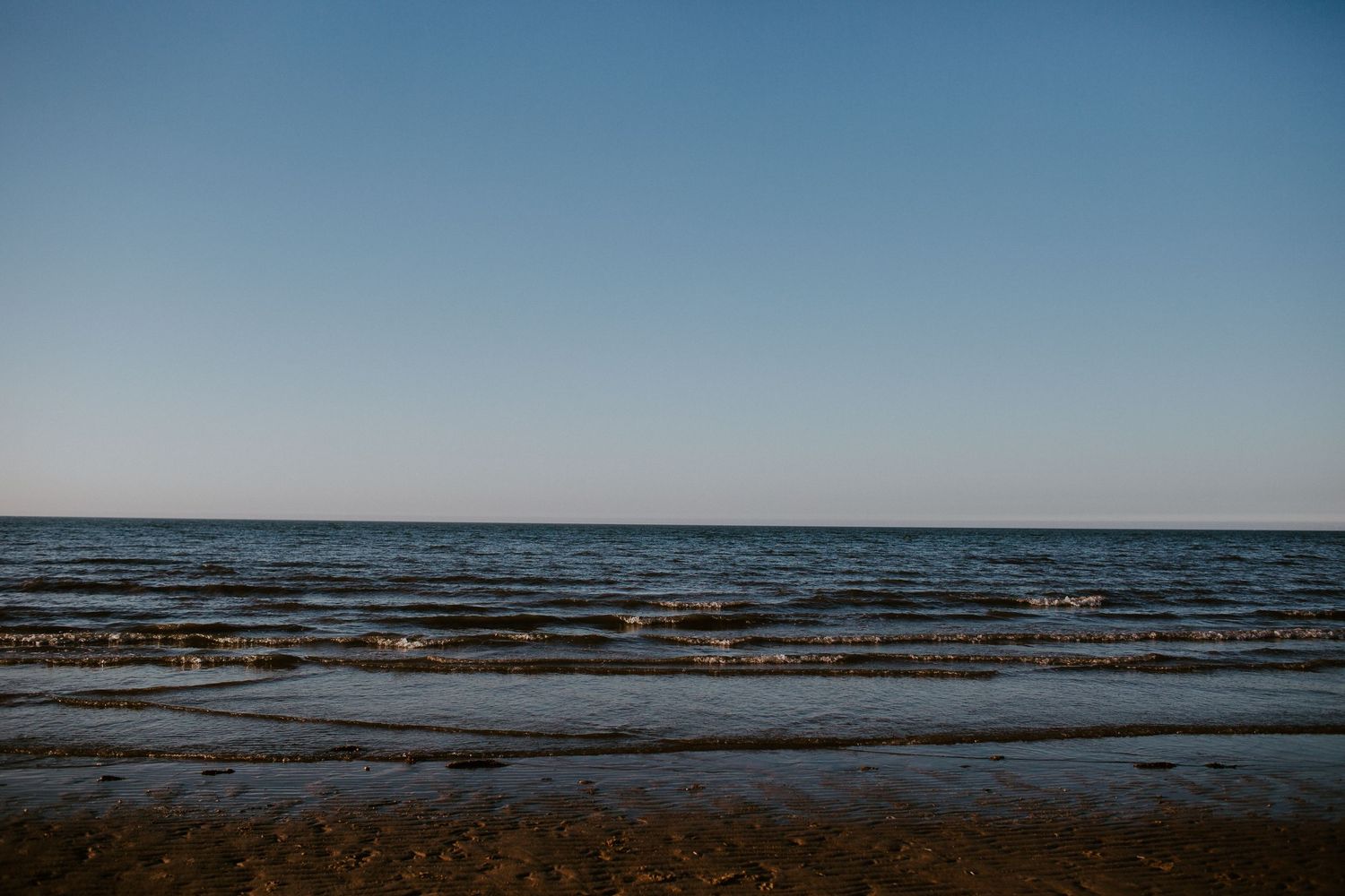 Calm ocean shoreline with gentle waves, sandy beach, and clear blue sky at dusk.