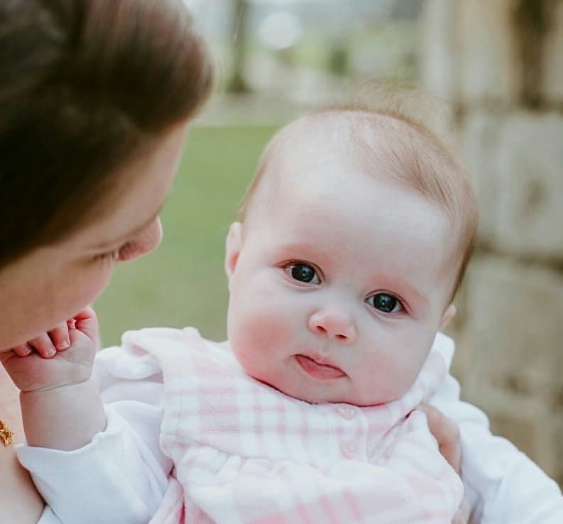 A baby in pink clothing smiles while being held against a soft background.