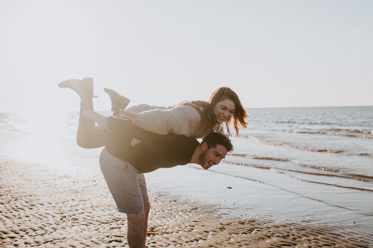 A couple playfully enjoys a sunny beach, with a man carrying a laughing woman on his back near the ocean.