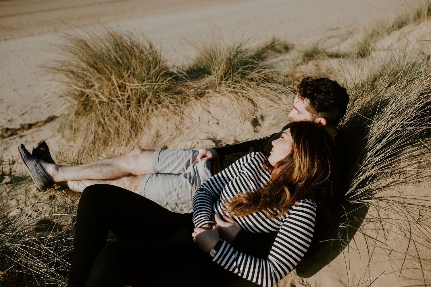 Couple relaxing together on sandy beach dunes surrounded by tall grass on a sunny day.