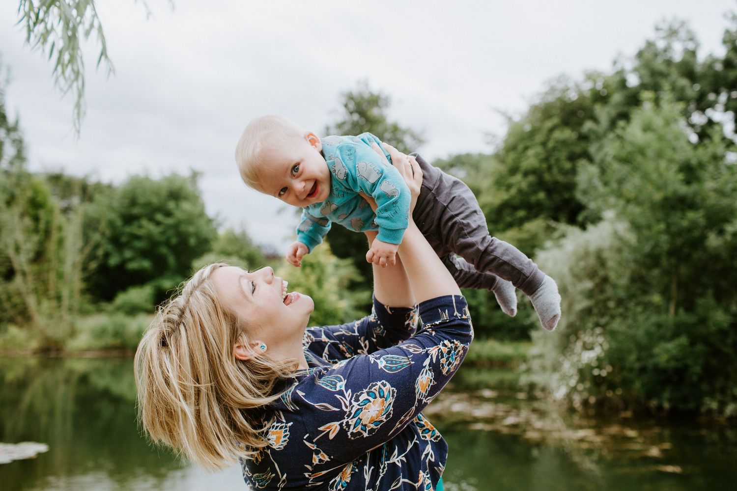 A joyful mother lifts her smiling baby outdoors near a lush green pond on a cloudy day.