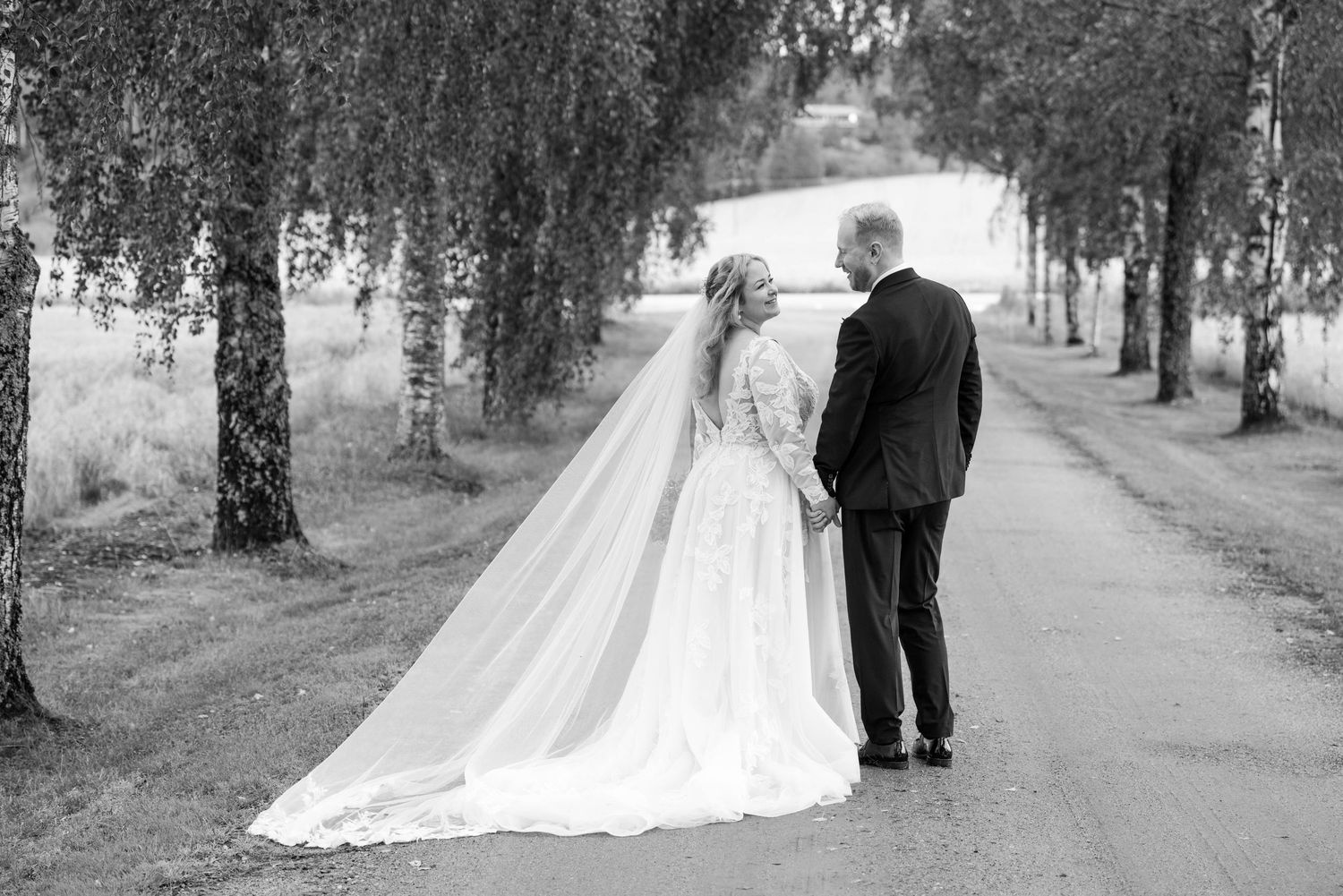 A black and white wedding portrait shows a couple on a tree-lined path in elegant attire.