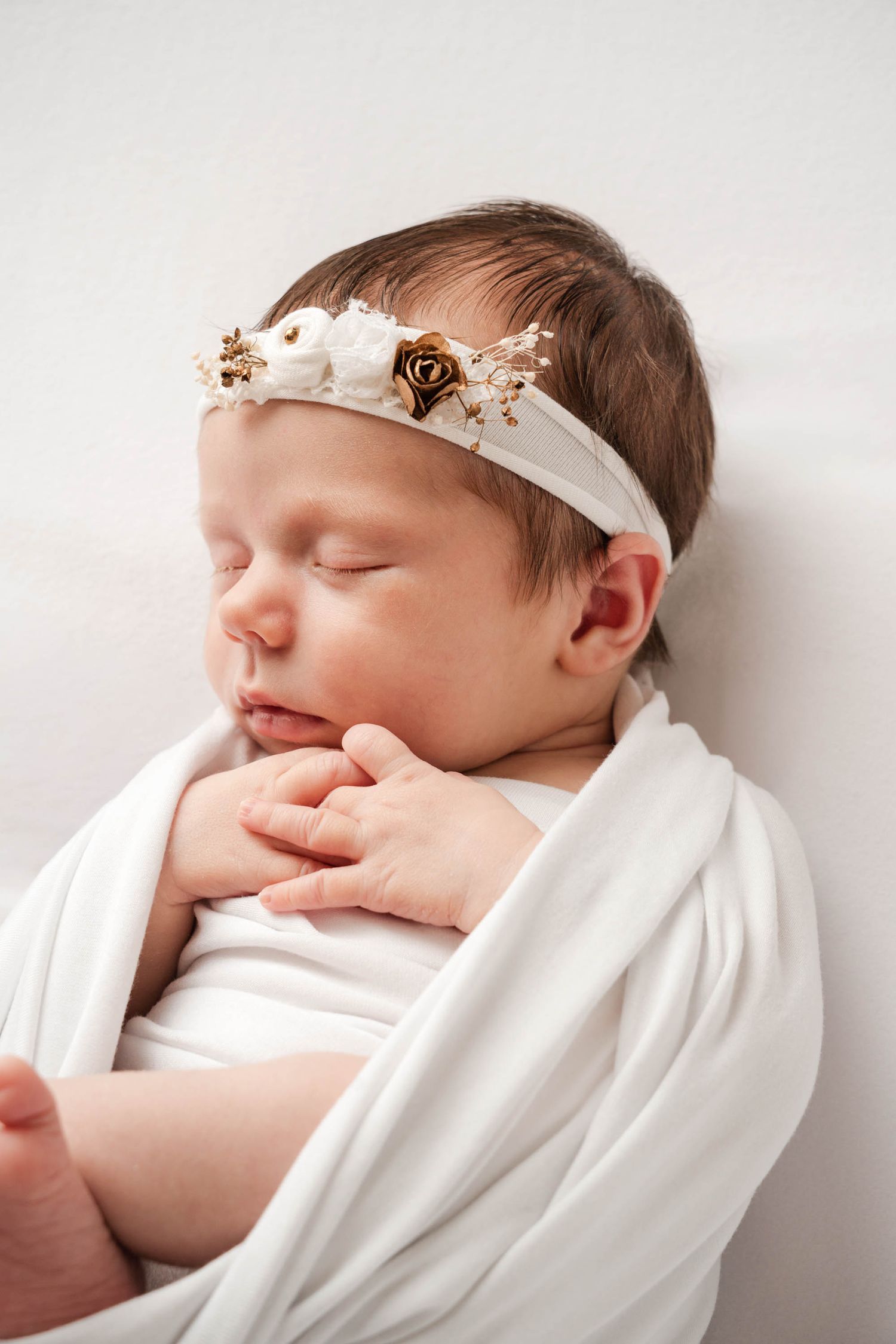 A sleeping newborn baby swaddled in white fabric with a delicate headband rests peacefully against a light background.
