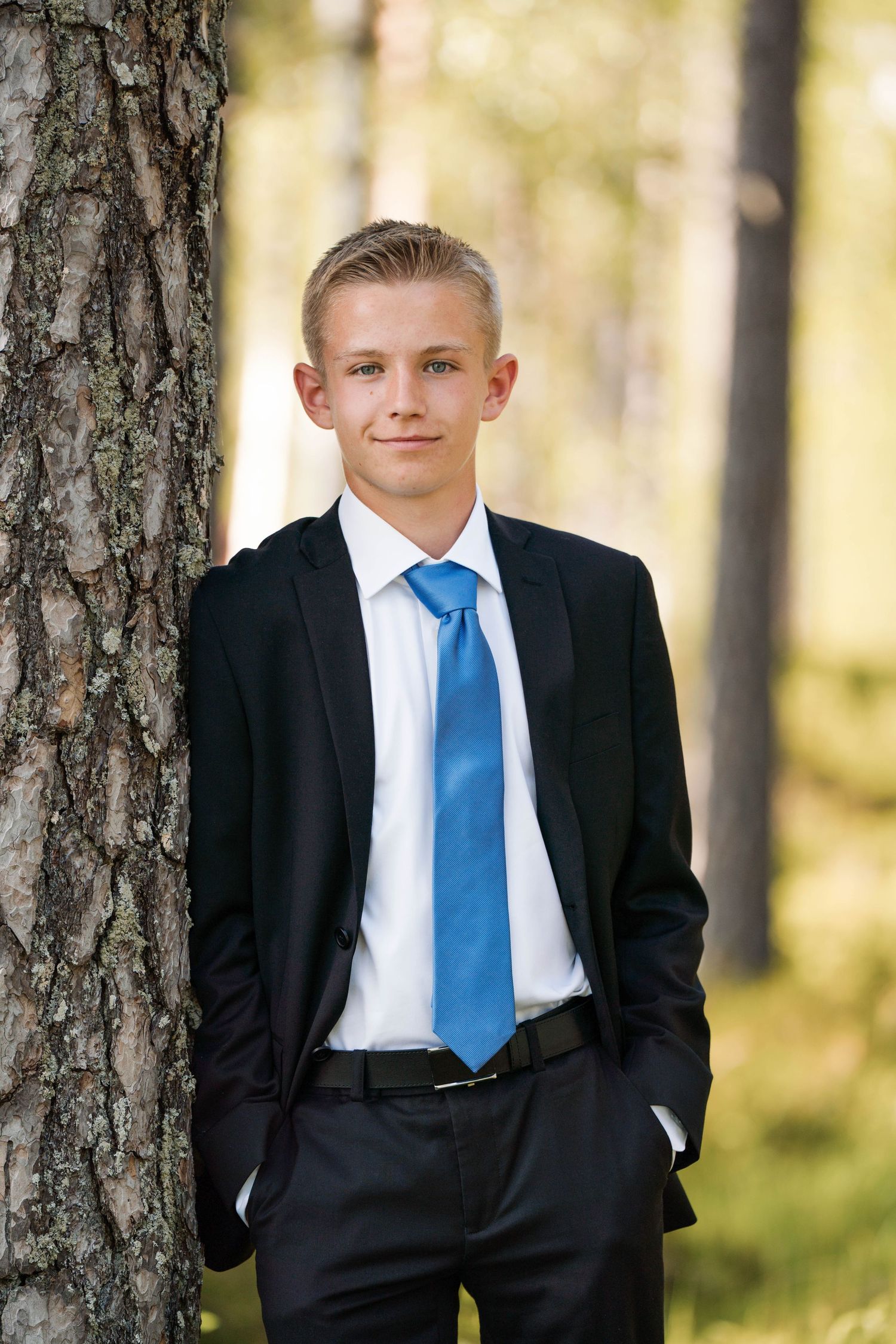 A formal portrait of a young person in a suit and blue tie leaning against a tree.