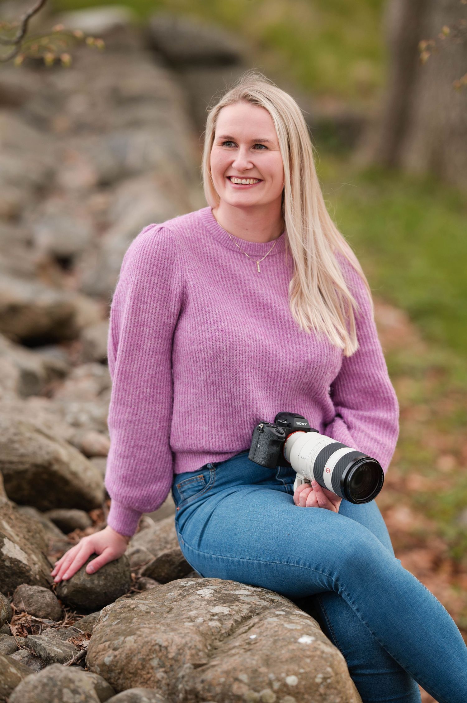 Woman in purple sweater sitting on rocks while holding camera gear.