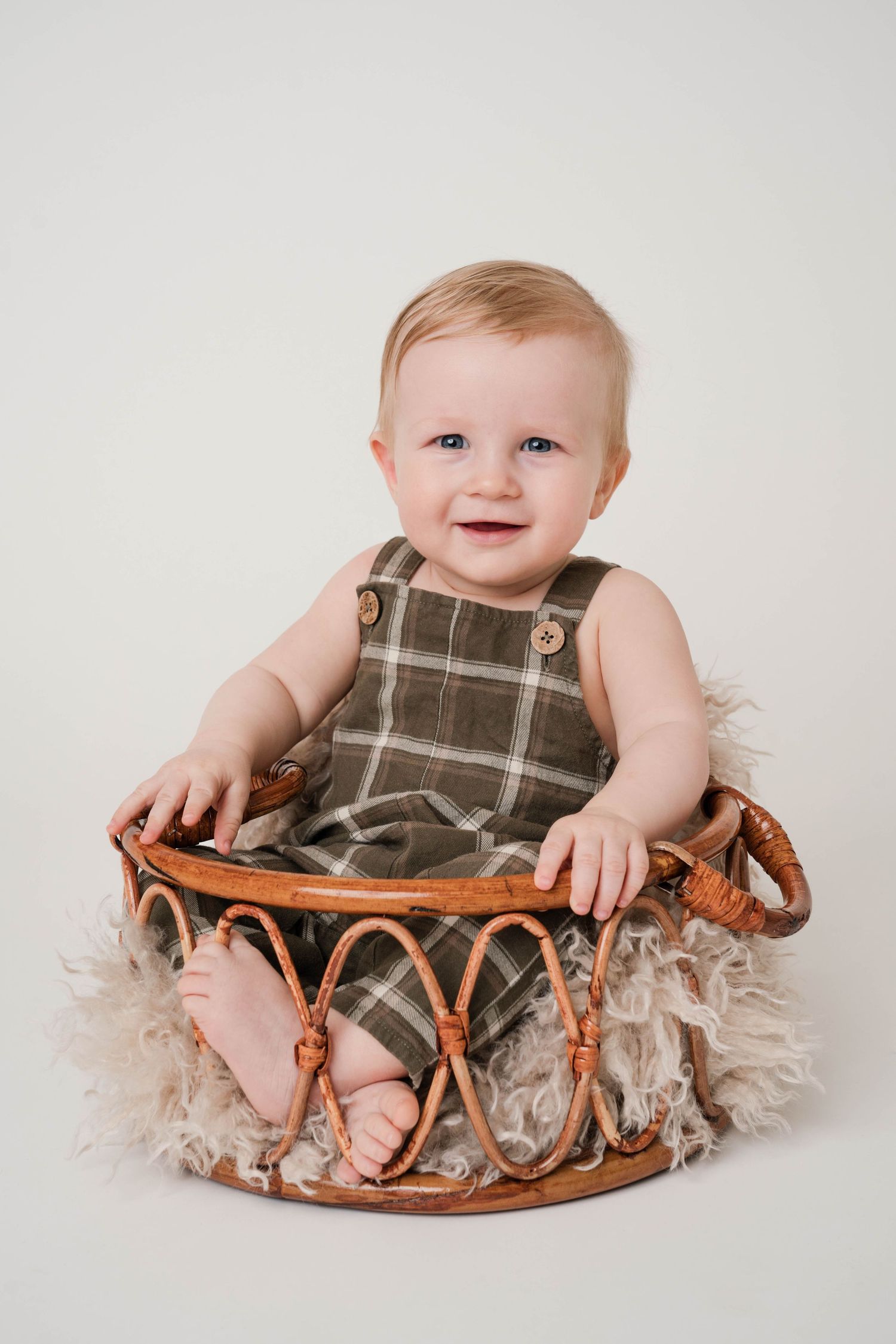 Smiling baby in plaid overalls sits in decorative basket with textured fabric.