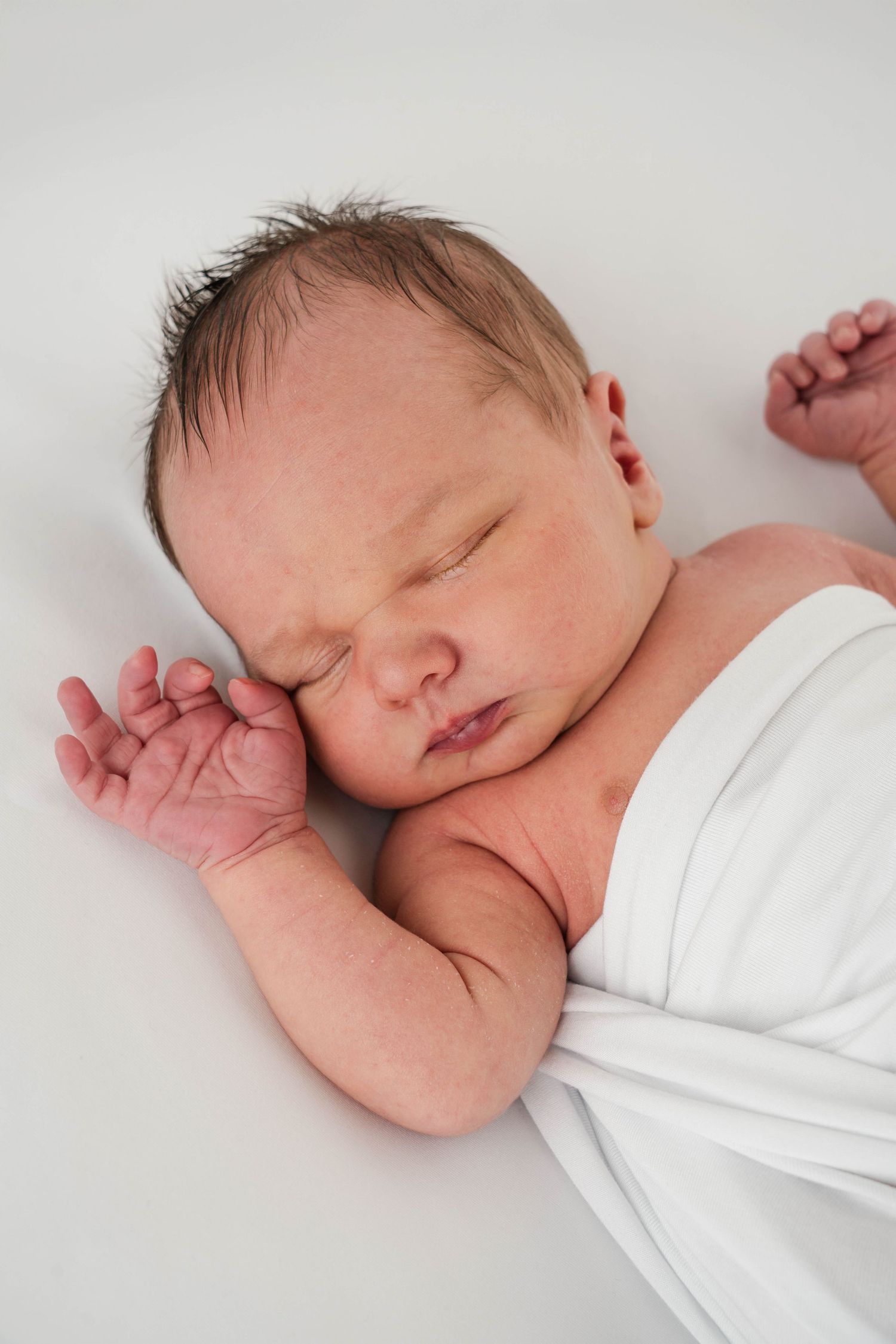 A sleeping newborn baby wearing white clothing rests peacefully against a light background.