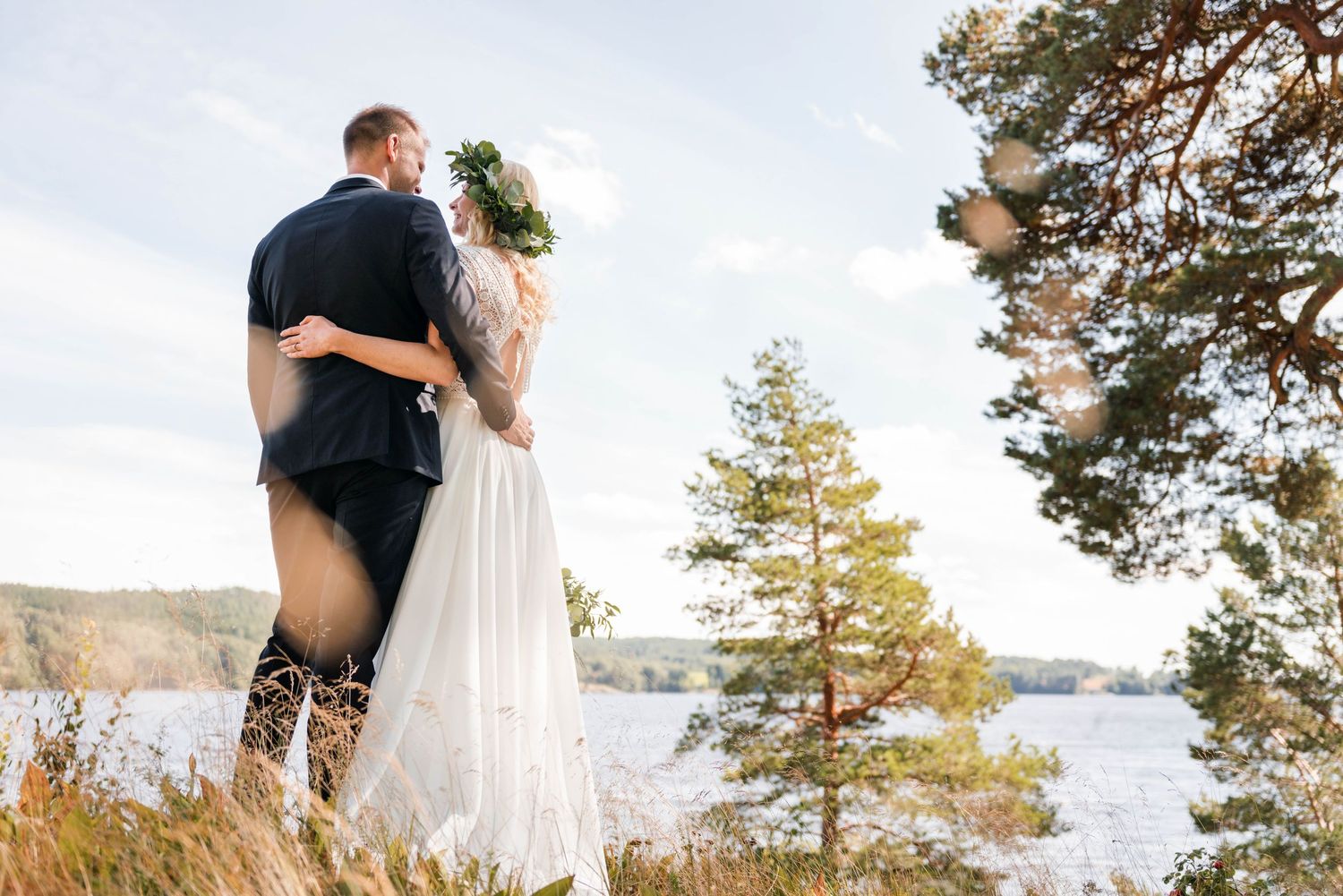 A couple in wedding attire embrace while standing on a rocky shoreline with pine trees and a lake in the background.