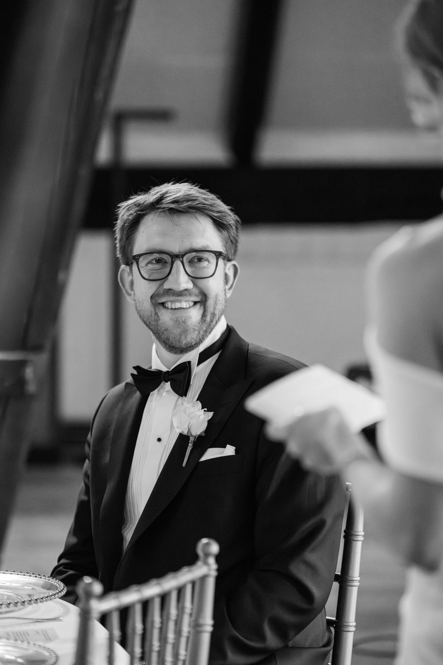 A groom in a black tuxedo and glasses smiles during a wedding reception.