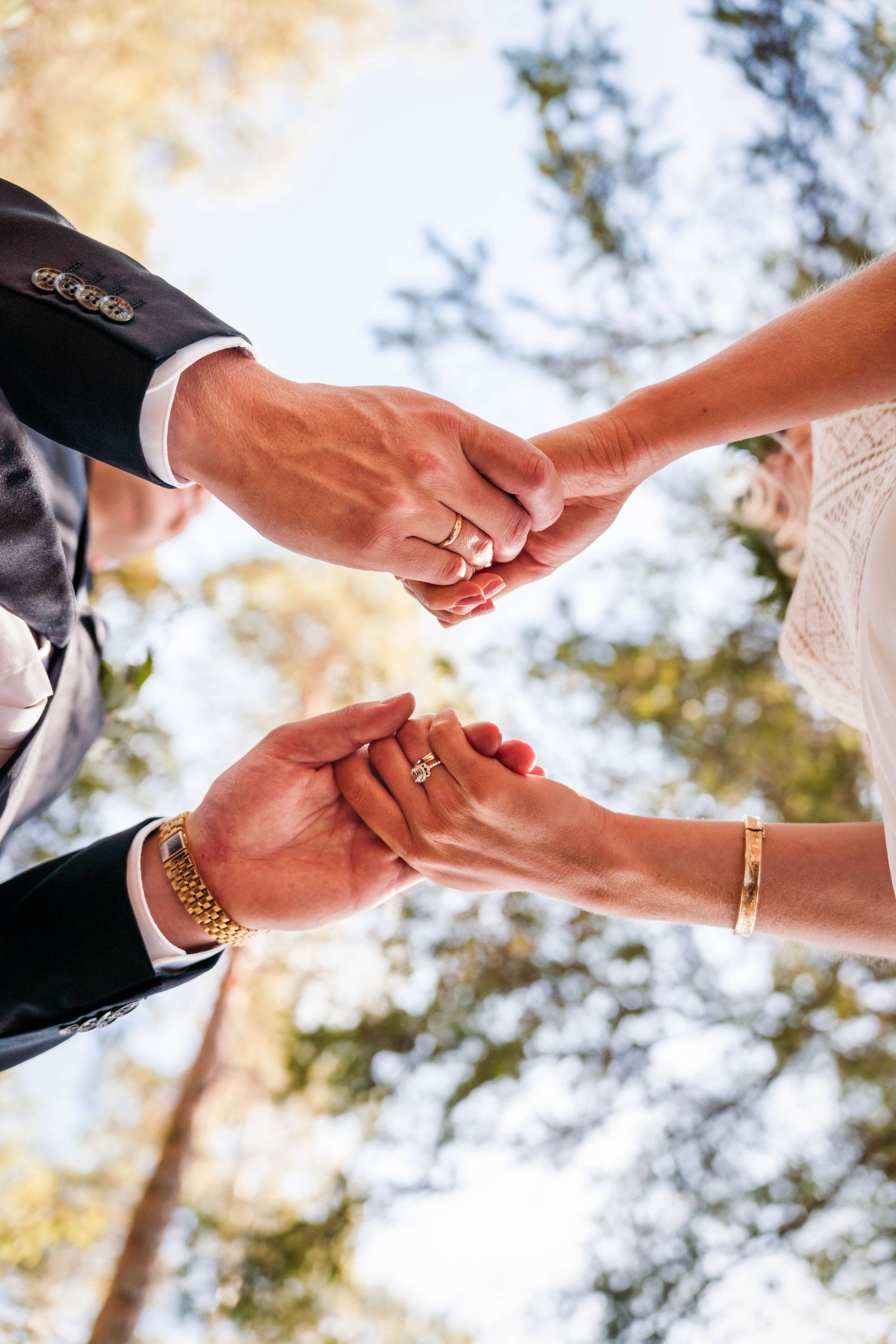 Close up of newlyweds holding hands showing wedding rings and jewelry against sunlit forest background.