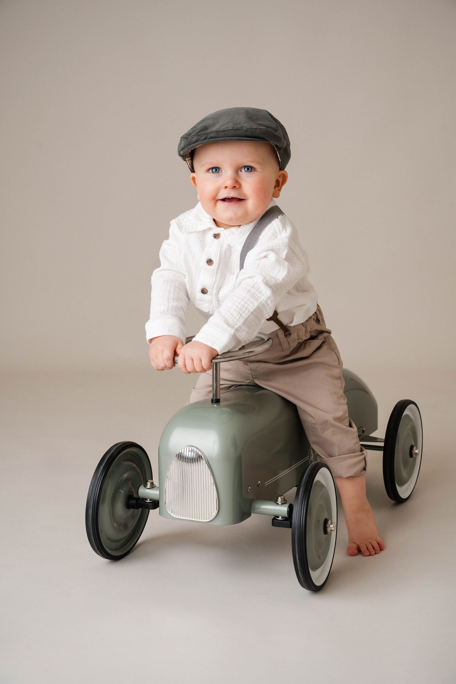 Smiling baby boy in vintage flat cap, white shirt, suspenders, riding a retro green toy car on a neutral background.