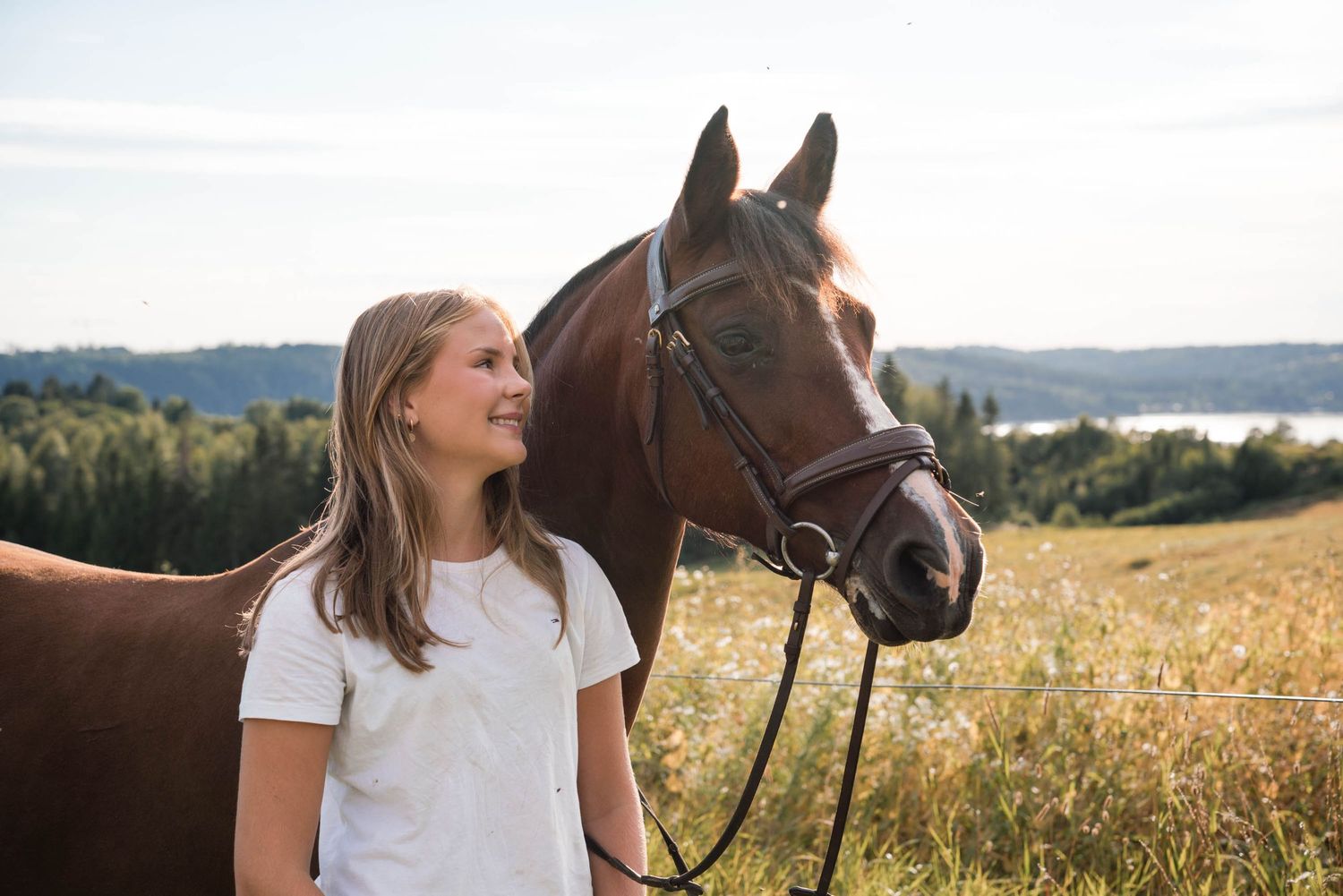 A woman in a white t-shirt stands next to a brown horse against a scenic countryside backdrop.