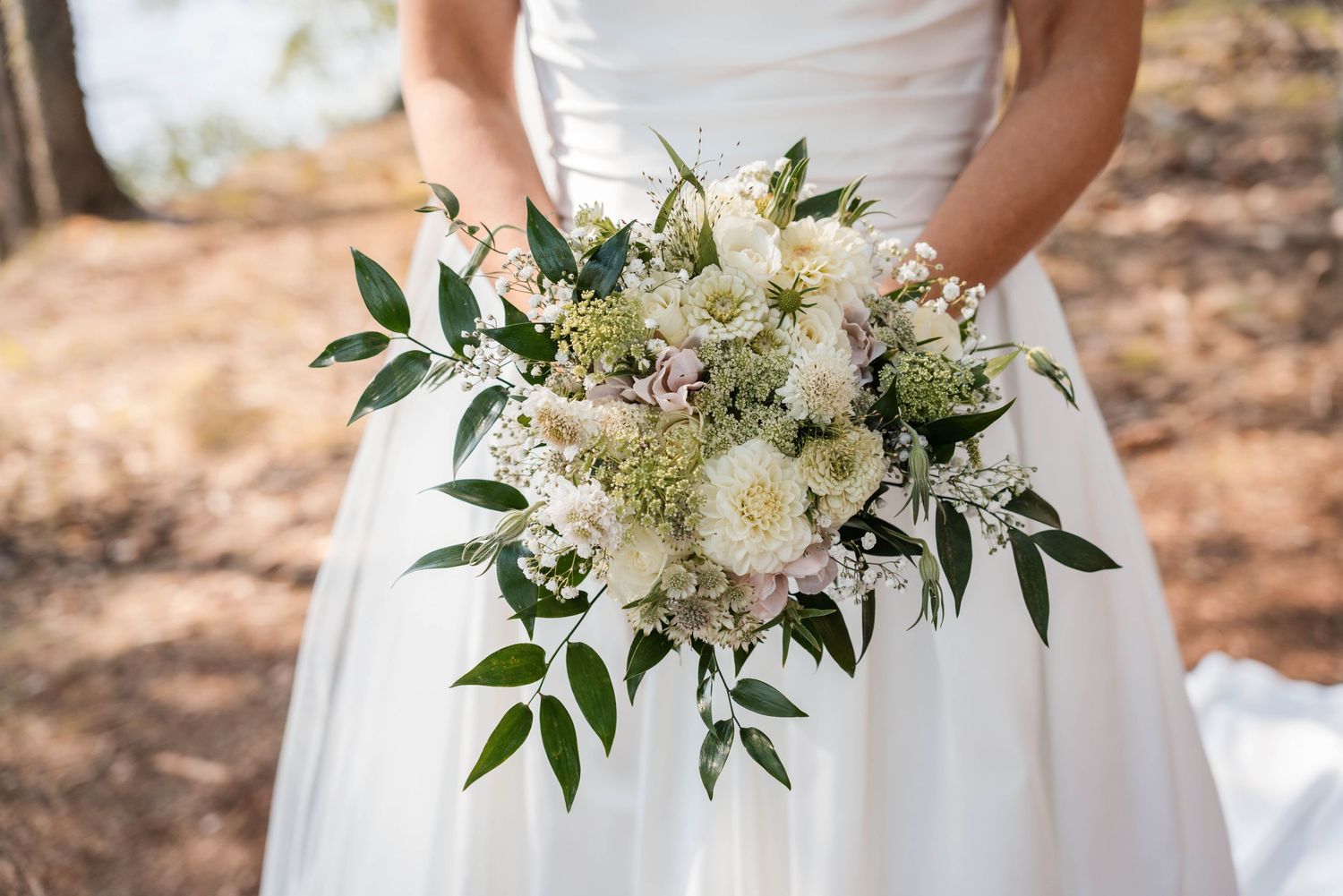 Elegant white bridal bouquet with lilies and greenery held against a natural outdoor backdrop.
