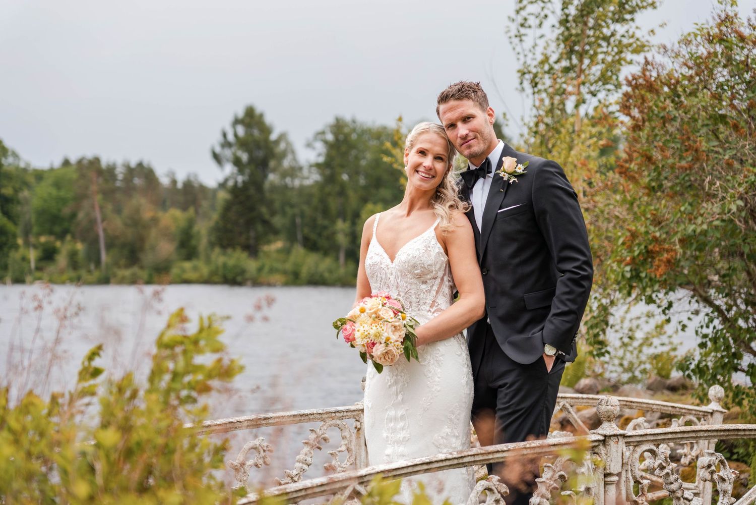 Romantic wedding portrait of couple by lakeside with autumn foliage and bouquet.