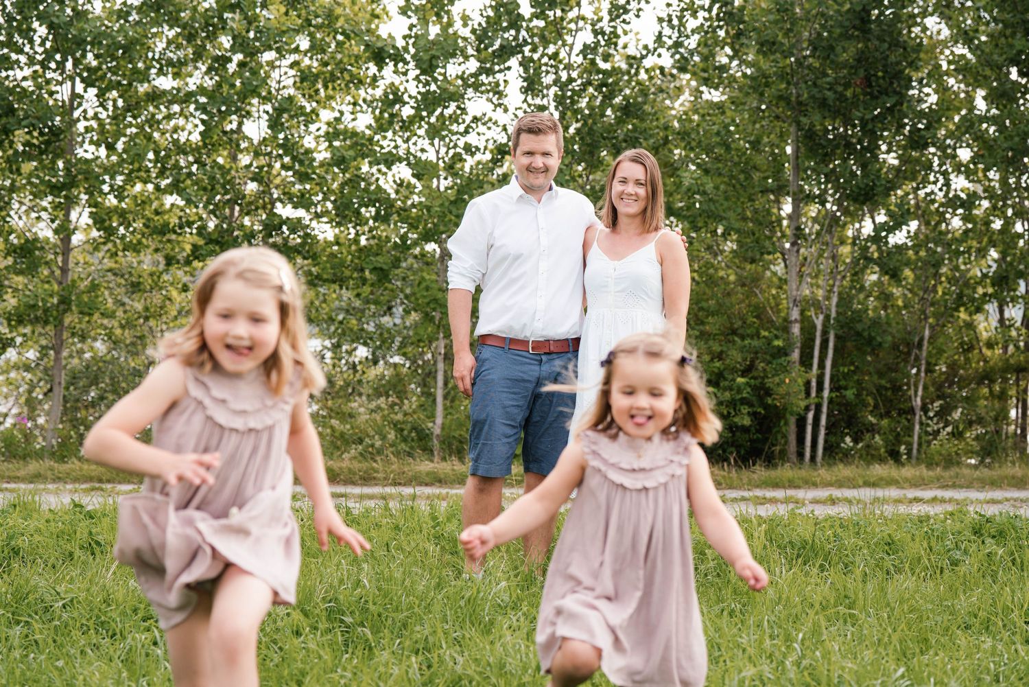 A playful family photo outdoors with two children running in matching pink dresses against a backdrop of birch trees.